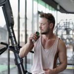 Muscular young sportsman in casual sportswear drinking fresh energy beverage near window while resting after intense workout in modern fitness club