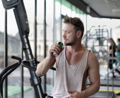 Muscular young sportsman in casual sportswear drinking fresh energy beverage near window while resting after intense workout in modern fitness club
