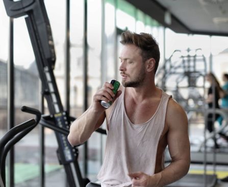 Muscular young sportsman in casual sportswear drinking fresh energy beverage near window while resting after intense workout in modern fitness club